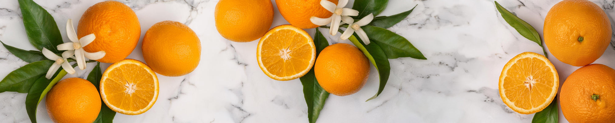 Fresh whole and sliced oranges arranged with green leaves and small white blossoms on a marble surface.