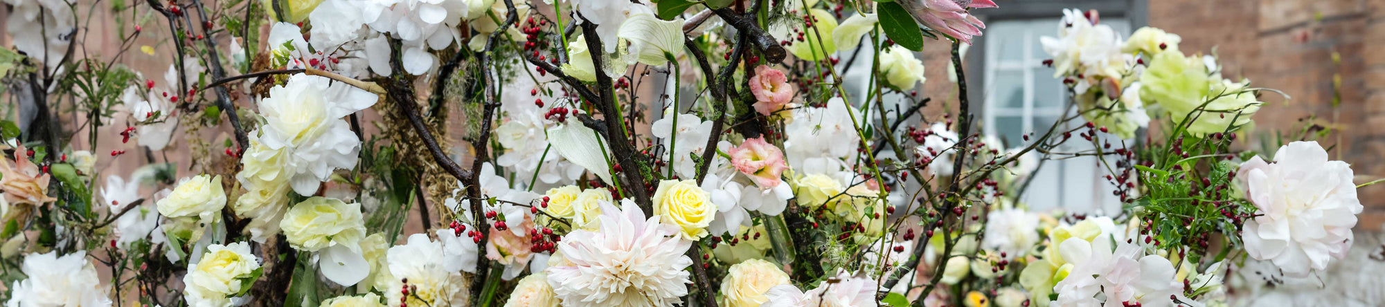 A lush floral arrangement featuring white, yellow, and pink flowers intertwined with green foliage and red berries, set against a blurred background of a brick building and a white window.